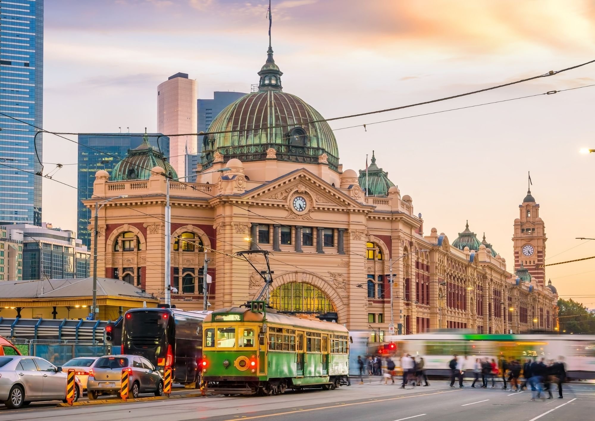 Flinders Street Station in Melbourne at sunset, with a green heritage tram, city traffic and pedestrians in the foreground.