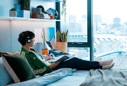 A young person relaxes in bed, headphones on, reading a book in a modern apartment with a city view.
