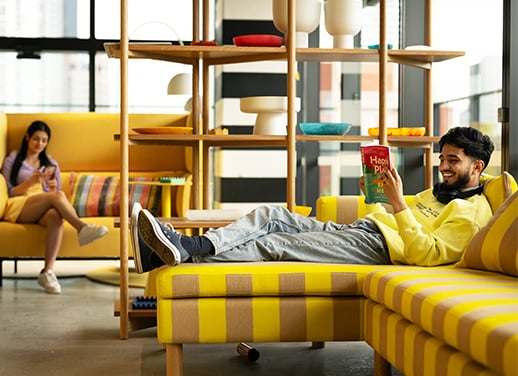 A man relaxes on a yellow couch reading while a woman sits on another yellow couch using her phone.